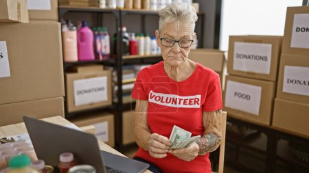 Téléchargez les photos : Femme âgée aux cheveux gris dévouée bénévole au centre communautaire de bienfaisance, comptant méticuleusement les dollars sur un ordinateur portable au milieu d'un tourbillon de dons. - en image libre de droit