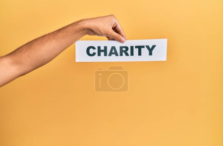 Téléchargez les photos : Hand of caucasian man holding paper with charity word over isolated yellow background - en image libre de droit