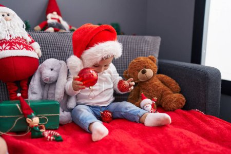 Téléchargez les photos : Adorable bébé caucasien jouant avec boule de décoration de Noël assis sur le canapé à la maison - en image libre de droit
