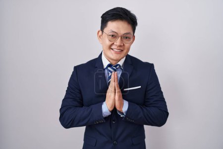 Téléchargez les photos : Jeune homme asiatique portant costume d'affaires et cravate priant avec les mains ensemble demandant pardon souriant confiant. - en image libre de droit