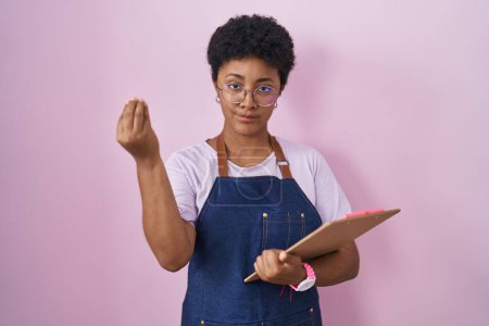 Téléchargez les photos : Jeune femme afro-américaine portant tablier de serveuse professionnel tenant presse-papiers faisant geste italien avec la main et les doigts expression confiante - en image libre de droit