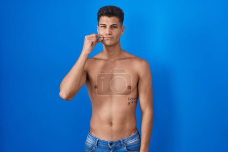 Foto de Joven hombre hispano de pie sin camisa sobre fondo azul boca y labios cerrados como cremallera con los dedos. secreto y silencioso, tabú hablando - Imagen libre de derechos