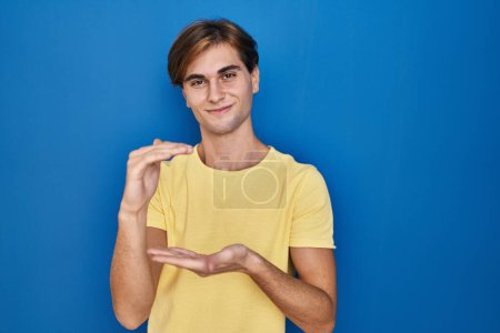 Photo for Young man standing over blue background gesturing with hands showing big and large size sign, measure symbol. smiling looking at the camera. measuring concept. - Royalty Free Image