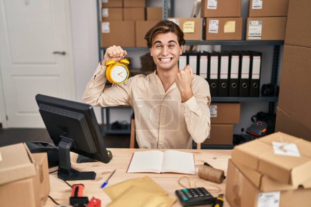 Photo for Young man working at small business ecommerce holding alarm clock screaming proud, celebrating victory and success very excited with raised arms - Royalty Free Image