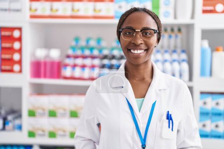 Foto de Farmacéutica mujer afroamericana sonriendo confiado de pie en la farmacia - Imagen libre de derechos