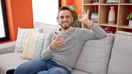 Photo for Hispanic man using smartphone doing ok gesture at home - Royalty Free Image