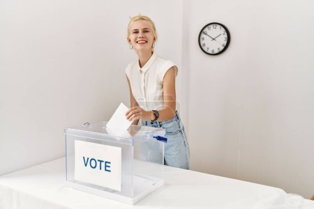 Foto de Joven rubia sonriendo confiada votando en colegio electoral - Imagen libre de derechos