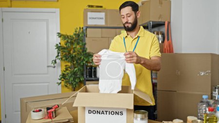 Téléchargez les photos : Jeune homme hispanique volontaire emballant des vêtements sur une boîte en carton pour faire un don au centre de bienfaisance - en image libre de droit