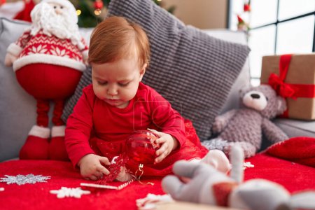 Téléchargez les photos : Adorable rousse tout-petit tenant boule de décoration de Noël assis sur le canapé à la maison - en image libre de droit