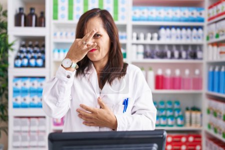 Téléchargez les photos : Femme brune d'âge moyen travaillant à la pharmacie droguerie sentant quelque chose de puant et dégoûtant, odeur intolérable, retenant la respiration avec les doigts sur le nez. mauvaise odeur - en image libre de droit