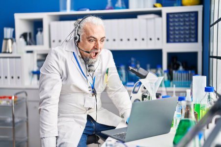 Photo for Middle age man with grey hair working at scientist laboratory doing video call scared and amazed with open mouth for surprise, disbelief face - Royalty Free Image