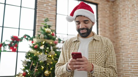Téléchargez les photos : Jeune homme hispanique célébrant Noël en utilisant un smartphone à la maison - en image libre de droit