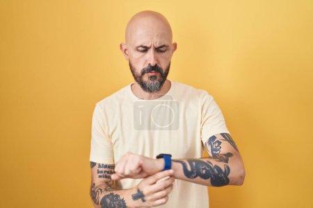 Foto de Hombre hispano con tatuajes de pie sobre fondo amarillo comprobando la hora en el reloj de pulsera, relajado y seguro - Imagen libre de derechos