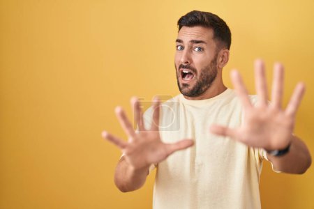 Photo for Handsome hispanic man standing over yellow background afraid and terrified with fear expression stop gesture with hands, shouting in shock. panic concept. - Royalty Free Image