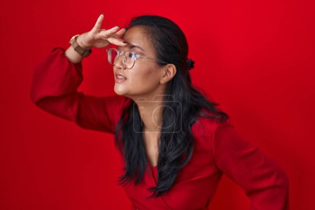 Photo for Asian young woman standing over red background very happy and smiling looking far away with hand over head. searching concept. - Royalty Free Image