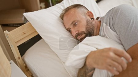 Téléchargez les photos : Beau jeune homme couché sur un lit confortable, montrant une expression sérieuse, reflétant un mode de vie concentré dans une chambre bien éclairée - en image libre de droit