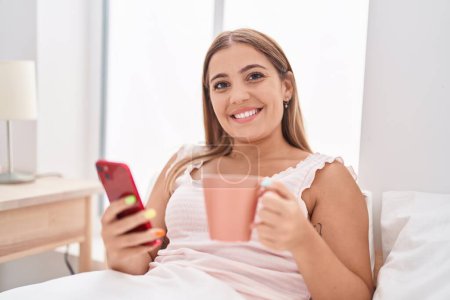 Téléchargez les photos : Young beautiful hispanic woman using smartphone drinking coffee at bedroom - en image libre de droit
