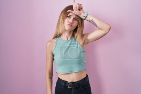 Foto de Blonde caucasian woman standing over pink background making fun of people with fingers on forehead doing loser gesture mocking and insulting. - Imagen libre de derechos