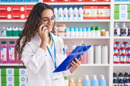 Téléchargez les photos : Jeune belle pharmacienne hispanique parlant sur smartphone document de lecture à la pharmacie - en image libre de droit