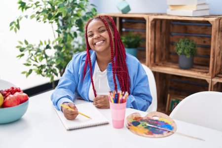 Téléchargez les photos : Femme artiste afro-américaine souriant dessin confiant sur ordinateur portable à la maison - en image libre de droit