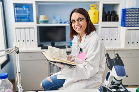 Téléchargez les photos : Jeune belle femme hispanique scientifique souriant livre de lecture confiant au laboratoire - en image libre de droit