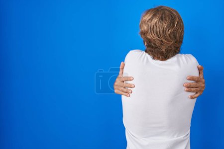 Téléchargez les photos : Homme d'âge moyen debout sur fond bleu se serrant dans ses bras heureux et positif à partir de l'arrière. amour de soi et soins de soi - en image libre de droit