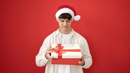 Téléchargez les photos : Jeune homme hispanique portant chapeau de Noël déballage cadeau à la recherche bouleversé sur fond rouge isolé - en image libre de droit