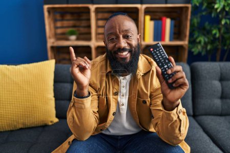 Foto de Hombre afroamericano sosteniendo el control remoto de la televisión sonriendo feliz señalando con la mano y el dedo hacia un lado - Imagen libre de derechos