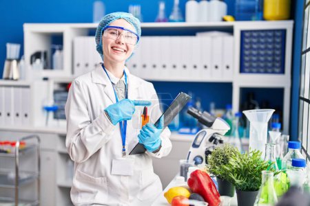 Foto de Hermosa mujer trabajando en el laboratorio científico con la comida sonriendo feliz señalando con la mano y el dedo - Imagen libre de derechos