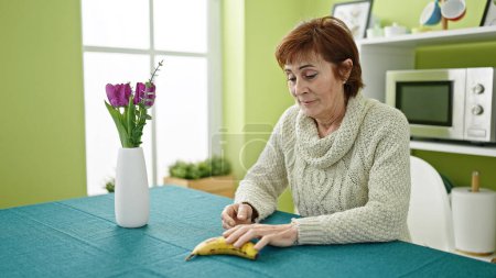 Téléchargez les photos : Mature hispanique femme assise sur la table manger une banane à la salle à manger - en image libre de droit