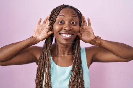 Foto de Mujer afroamericana de pie sobre fondo rosa tratando de escuchar ambos gestos de las manos en la oreja, curioso por chismes. problema auditivo, sordo - Imagen libre de derechos
