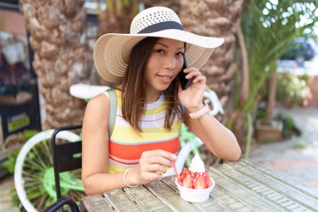 Photo for Young asian woman tourist eating ice cream talking on smartphone at coffee shop terrace - Royalty Free Image