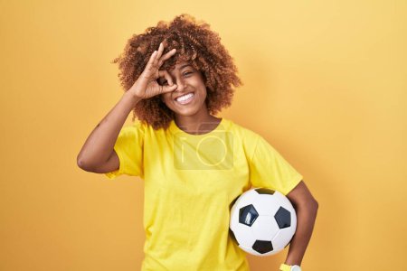 Foto de Mujer hispana joven con el pelo rizado sosteniendo pelota de fútbol sonriendo feliz haciendo signo bien con la mano en el ojo mirando a través de los dedos - Imagen libre de derechos