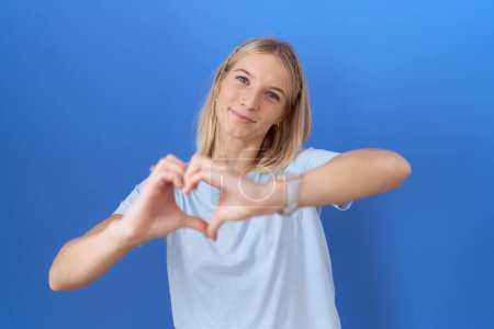 Téléchargez les photos : Jeune femme caucasienne portant un t-shirt bleu décontracté souriant en amour faisant forme de symbole de coeur avec les mains. concept romantique. - en image libre de droit