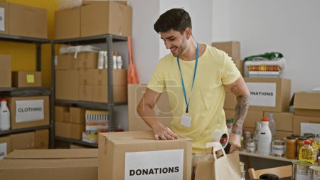 Téléchargez les photos : Jeune homme hispanique bénévole souriant confiant emballage dons boîte en carton au centre de charité - en image libre de droit