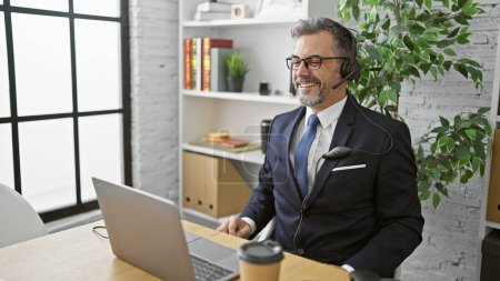 Photo for Smiling young hispanic man, a grey-haired business worker, buzzes with success as he handles business on his laptop, headphones on, in a lively office. - Royalty Free Image