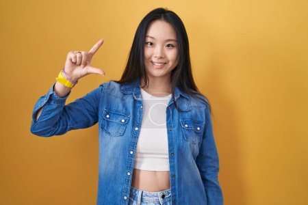 Téléchargez les photos : Jeune femme asiatique debout sur fond jaune geste souriant et confiant avec la main faisant signe de petite taille avec les doigts regardant et la caméra. concept de mesure. - en image libre de droit