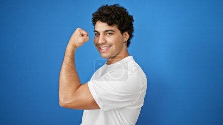 Téléchargez les photos : Jeune homme latin souriant confiant faire un geste fort avec le bras sur fond bleu isolé - en image libre de droit