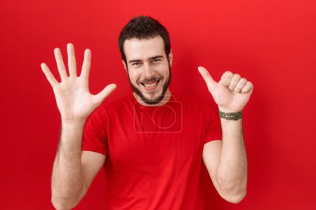 Téléchargez les photos : Jeune homme hispanique portant t-shirt rouge décontracté montrant et pointant vers le haut avec les doigts numéro six tout en souriant confiant et heureux. - en image libre de droit