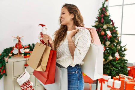 Téléchargez les photos : Young beautiful hispanic woman holding shopping bags standing by christmas tree at home - en image libre de droit