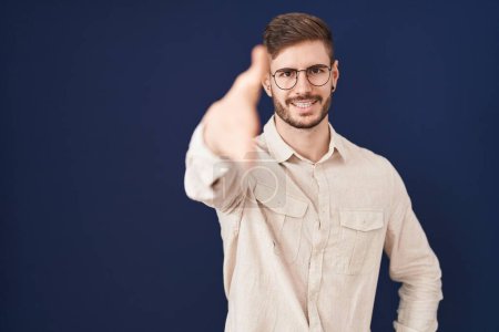 Photo for Hispanic man with beard standing over blue background smiling friendly offering handshake as greeting and welcoming. successful business. - Royalty Free Image