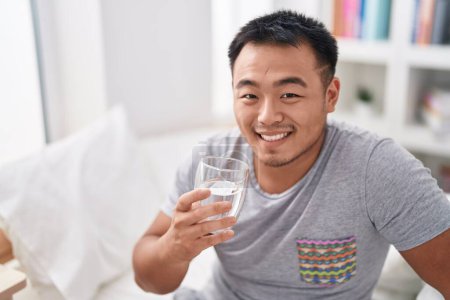 Photo for Young chinese man drinking glass of water sitting on bed at bedroom - Royalty Free Image