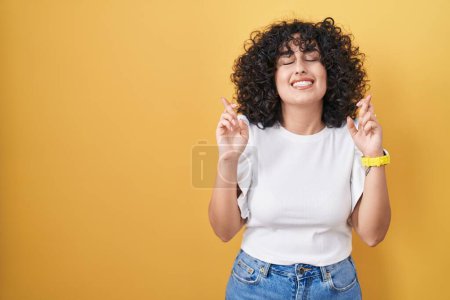 Téléchargez les photos : Jeune femme du Moyen-Orient debout sur fond jaune geste doigt croisé souriant avec espoir et les yeux fermés. chance et concept superstitieux. - en image libre de droit