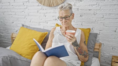 Foto de Senior mujer de pelo gris beber taza de libro de lectura de café en el dormitorio - Imagen libre de derechos
