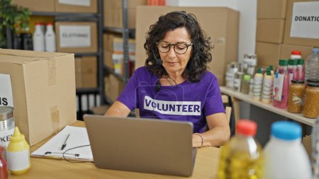Téléchargez les photos : Moyen Age hispanique femme bénévole en utilisant ordinateur portable et écouteurs au centre de charité - en image libre de droit
