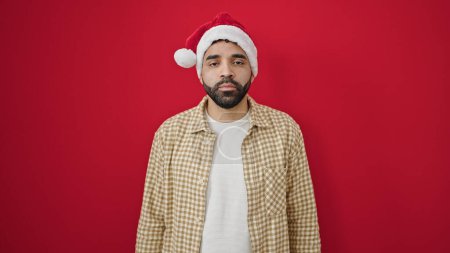 Photo for Young hispanic man standing with serious expression wearing christmas hat over isolated red background - Royalty Free Image