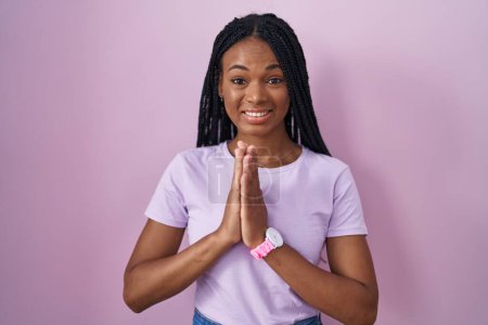 Foto de Mujer afroamericana con trenzas de pie sobre fondo rosa rezando con las manos juntas pidiendo perdón sonriendo confiado. - Imagen libre de derechos