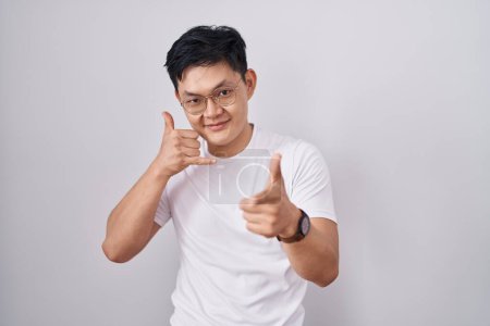 Téléchargez les photos : Jeune homme asiatique debout sur fond blanc souriant faire parler au téléphone geste et vous pointant du doigt. Appelle-moi. - en image libre de droit