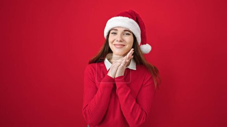 Foto de Mujer hispana joven sonriendo confiada usando sombrero de Navidad sobre fondo rojo aislado - Imagen libre de derechos