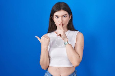 Téléchargez les photos : Young caucasian woman standing over blue background asking to be quiet with finger on lips pointing with hand to the side. silence and secret concept. - en image libre de droit